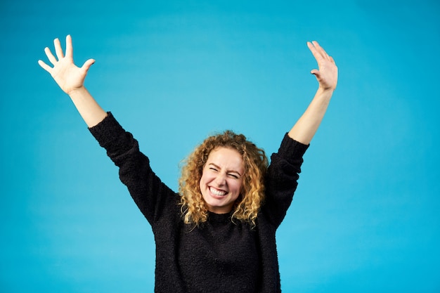 Donna riccia della giovane testarossa soddisfatta felice che celebra e che incoraggia un successo che spande le mani con gioia contro il blu