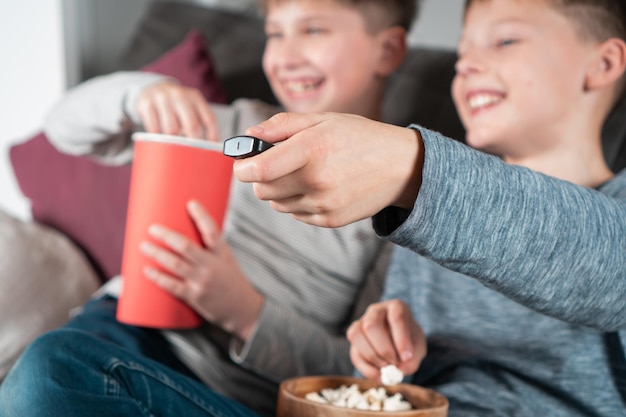 Happy school aged boys sit on sofa eat popcorn and watch TV Focus on outstretched hand holding TV remote control