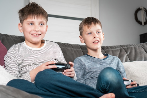 Happy school aged boys sit on sofa with game joysticks Children play videogames Interesting amusement competition