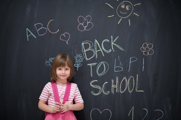 Bambina felice della scuola con lo zaino che scrive di nuovo a scuola sulla lavagna nera