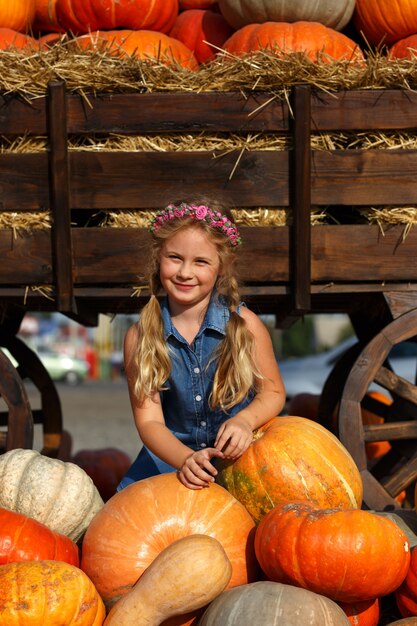 Happy school girl sitting between pumpkins at local farmer market in sunny autumn day.