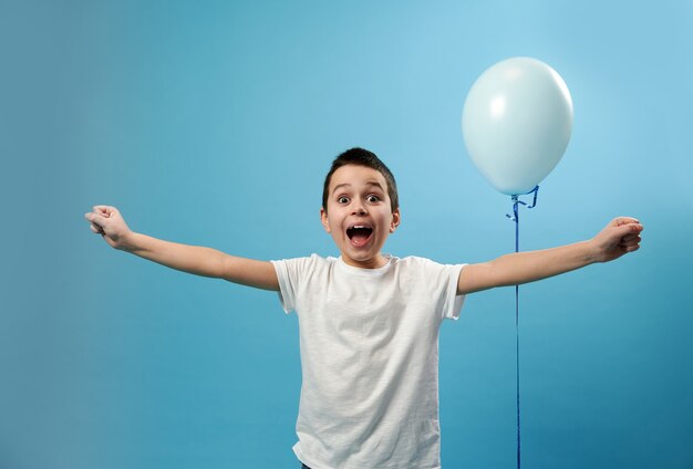 Happy schoolboy expressing happiness standing on blue surface near a blue balloon