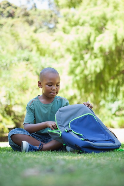 Happy schoolboy opening his schoolbag sitting on grass