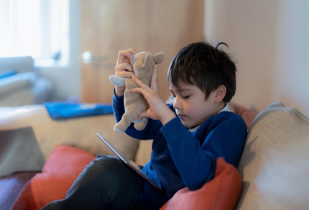 Happy schoolboy playing with dog toy and game online with friend on tabletKid using internet sending homework to the teacher Positive child sitting on sofa relaxing in living room in the morning