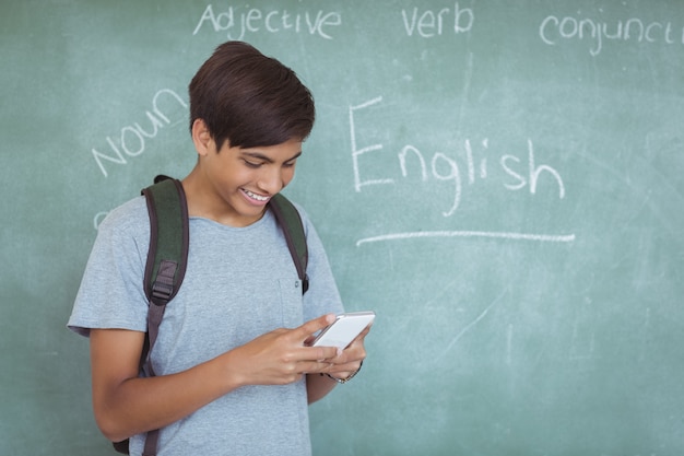 Happy schoolboy with backpack using mobile phone in classroom