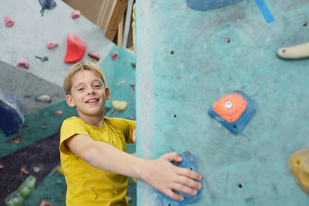 Happy schoolboy in yellow t-shirt holding by small rocks on climbing equipment while looking at you during training