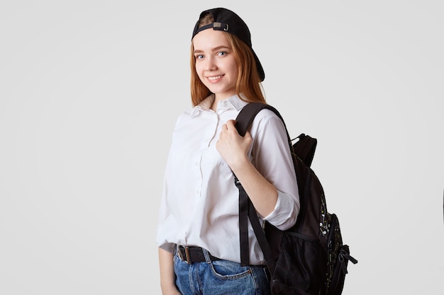 Happy schoolgirl in black cap and white shirt, carries rucksack with books, ready for starting new studying year, poses on white. College student returns back home from classes