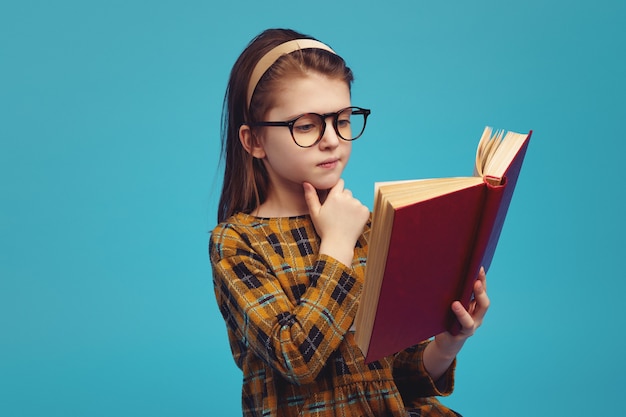 Happy schoolgirl in eyeglasses reading book and doing homework