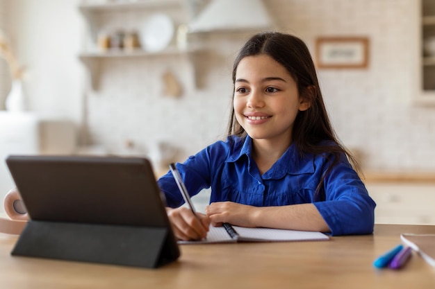 Photo happy schoolgirl looking at digital tablet taking notes at home