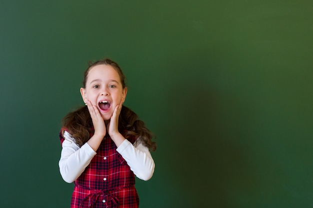 Ragazza prescolare della scolara felice in vestito dal plaid che sta nella classe vicino ad una lavagna verde.