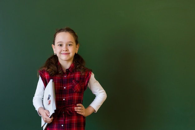 Happy schoolgirl preschool girl in plaid dress standing in class near a green blackboard. 