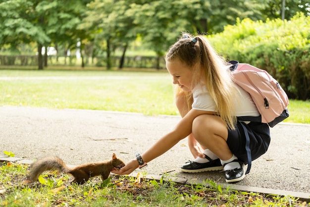 배낭을 메고 제복을 입은 행복한 여학생은 학교에 가는 길에 공원에서 다람쥐에게 먹이를 준다