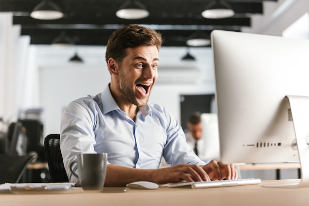 コンピューターを使用して幸せな叫び声のビジネスマンは、オフィスのテーブルのそばに座って喜ぶ