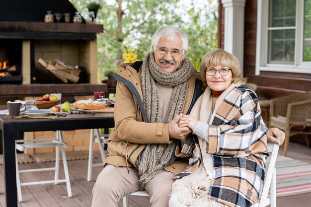 Happy senior affectionate grandparents in warm casualwear looking at you with smiles while sitting on chairs against served table by their house