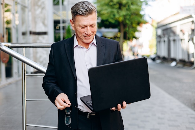 Фото Счастливый старший бизнесмен держа компьтер-книжку в его руке в городе внешнем и смотря экран.