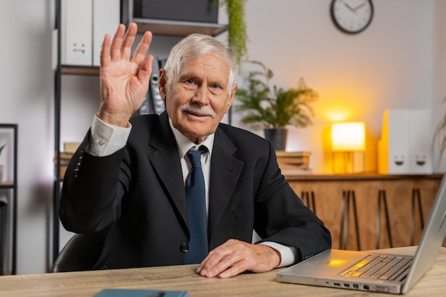 Happy senior businessman with laptop waves hand hi gesture greeting welcomes someone at home office