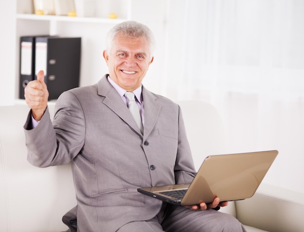 Счастливый старший бизнесмен работает на ноутбуке и показывает палец вверх.