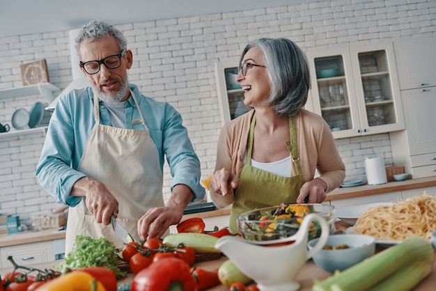 Happy senior couple in aprons preparing healthy dinner and smiling while spending time at home