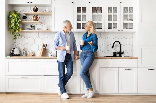 Happy senior couple drinking morning coffee and chatting in kitchen at home