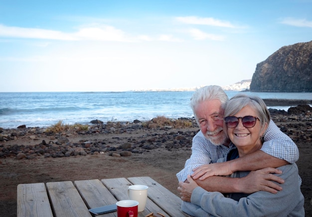 A happy senior couple embracing each other on the beach Sitting at a wooden table with two cups of coffee Two real people