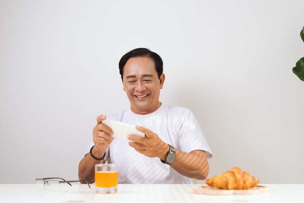 Happy senior couple having croissant breakfast at home and drinking orange juice, using smartphone.