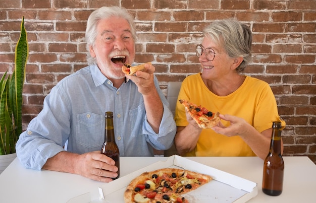 Happy senior couple having fun together eating a pizza Sitting at home table with beer and pizza Celebrating holiday in company