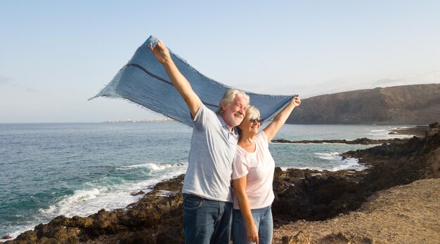 Photo happy senior couple holding textile while standing on rocky coastline