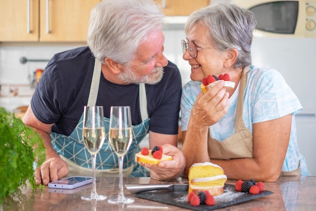 Happy senior couple in home kitchen looking in the eyes eating a handmade plumcake with berries