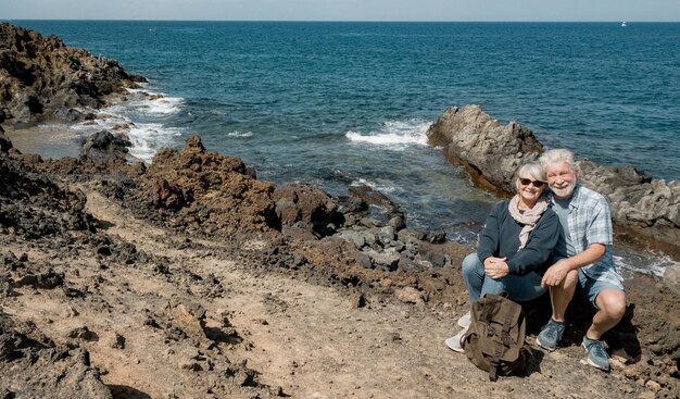 Happy senior couple in outdoors excursion sitting at the rock beach looking at the drone camera