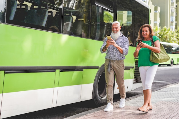 Photo happy senior couple using smartphones at bus station