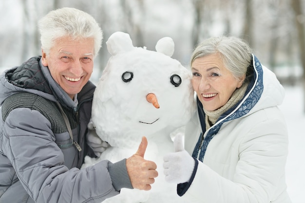 Happy senior couple with snowman in winter outdoors