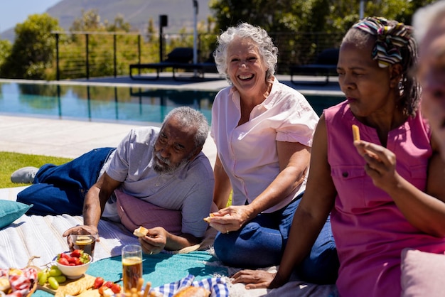 Photo happy senior diverse people sitting on blanket and having picnic in garden. senior lifestyle, friendship and relaxation, retirement, unaltered.