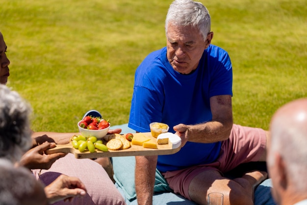 Happy senior diverse people sitting on blanket and having picnic in garden