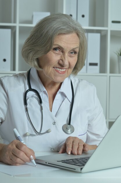 Happy senior doctor sitting at table with laptop in office