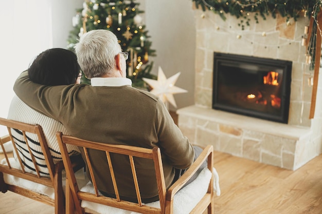 Happy senior family enjoying cozy fireplace Happy holidays Beautiful elderly couple hugging and relaxing in stylish christmas decorated living room Cozy winter