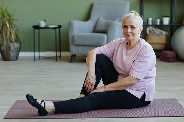 Happy senior female in sports clothes sitting on yoga mat on the floor of livingroom on background of armchair and small table