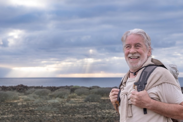 Photo happy senior grandfather enjoying excursion outdoors at sea holding backpack. horizon over water and dramatic sky with sunray