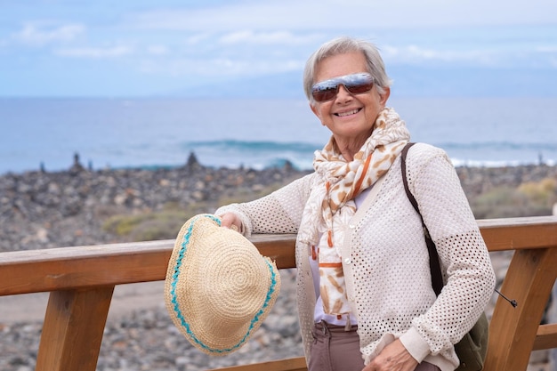 Happy senior gray haired woman with backpack walking along the sea beach with hat in hand enjoying vacation free time or retirement