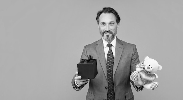 Happy senior grizzled man hold toy bear and box on grey background with copy space birthday