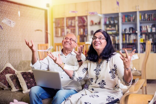 Happy Senior Indian asian couple having won lottery or investment earning in form of Money or paper currency falling from sky - raining concept