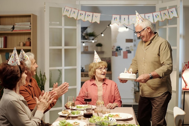 Felice uomo anziano che porta torta di compleanno fatta in casa per sua moglie alla festa