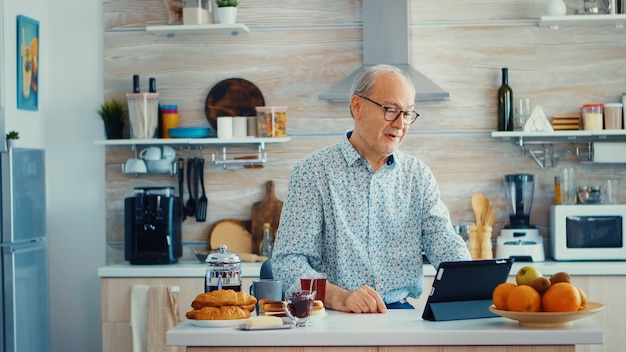 Happy senior man during breakfast in kitchen having a video\
conference with family. elderly person using internet online chat\
technology video webcam making a video call connection camera\
communicatio