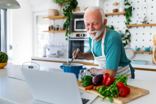 Happy senior man having fun cooking at home Elderly person preparing healthy lunch in modern kitchen looking at the receipt at his laptop