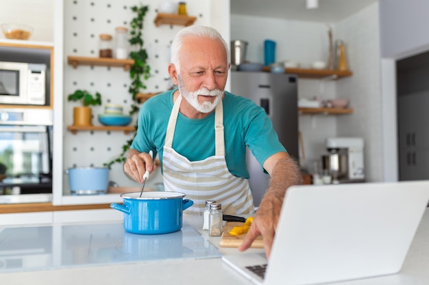 Happy senior man having fun cooking at home Elderly person preparing healthy lunch in modern kitchen looking at the receipt at his laptop