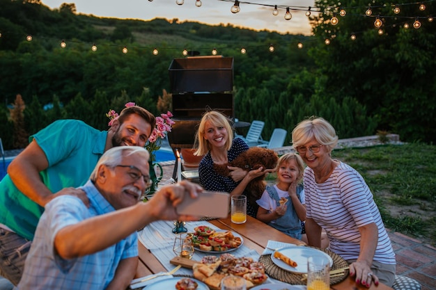 Foto felice uomo anziano e la sua famiglia allargata che prendono selfie durante la cena nel cortile