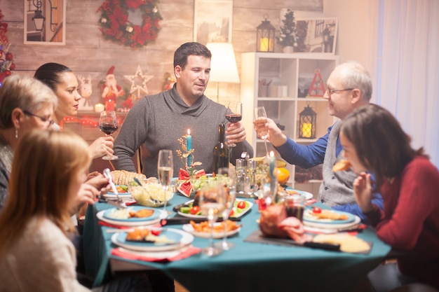 幸せな年配の男性と彼の息子は、クリスマスの家族の夕食時にグラスワインをチリンと鳴らします。