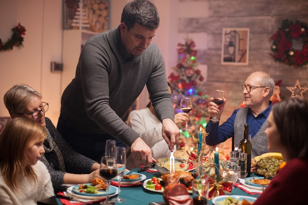 グラスワインを持っている幸せな年配の男性と彼の息子はクリスマスのお祝いで鶏肉をスライスします。