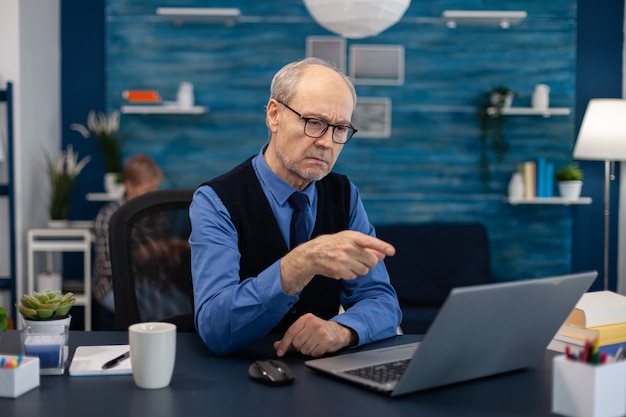 Happy senior man pointing at laptop during video conference. Cheerful pensioner with gray hair smiling at laptop webcam while chatting sitting at desk .