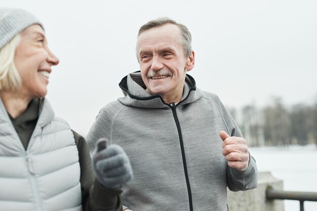 Happy senior man with mustache wearing gray sweatshirt talking to wife while they running together along riverside in winter