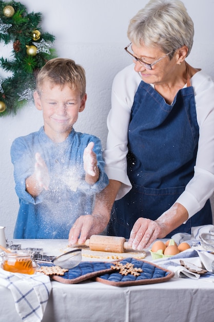 Happy senior mature woman, grandmother and young boy, grandson cooking, kneading dough, baking pie, cake,  cookies. Family time in the cozy kitchen. Seasonal winter Christmas activity at home.